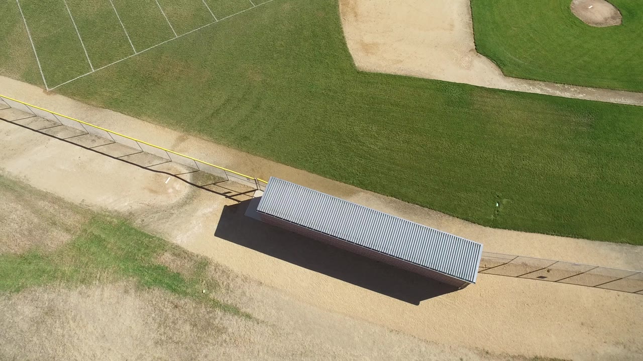 Aerial view of a baseball field corner with a small mound area, green grass, tan dirt, fencing, and a roofed shelter.