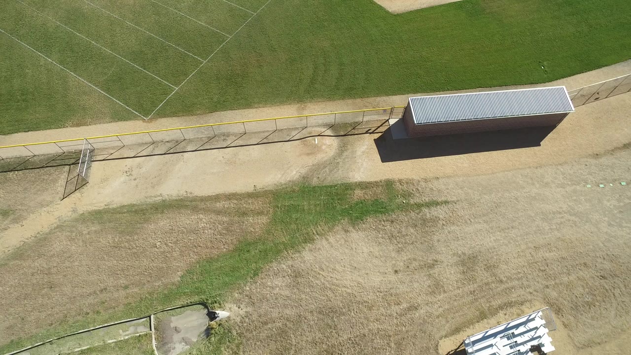 High aerial view over an empty baseball field in Hidden Lake, Colorado, with green outfield grass, tan dirt, a fence line, a roofed dugout, and no visible people.