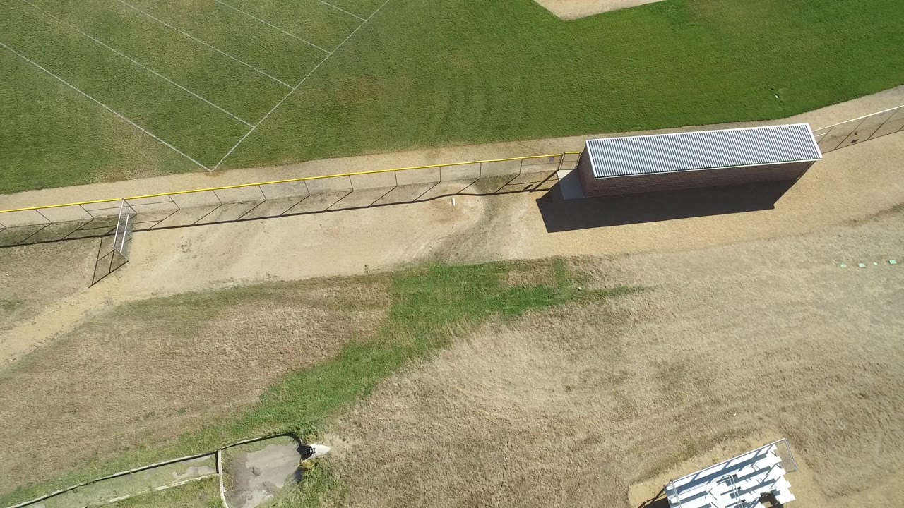 Overhead aerial frame in Hidden Lake, Colorado, showing the baseball field fence, dugout roof, patchy grass, tan dirt paths, and no visible people.