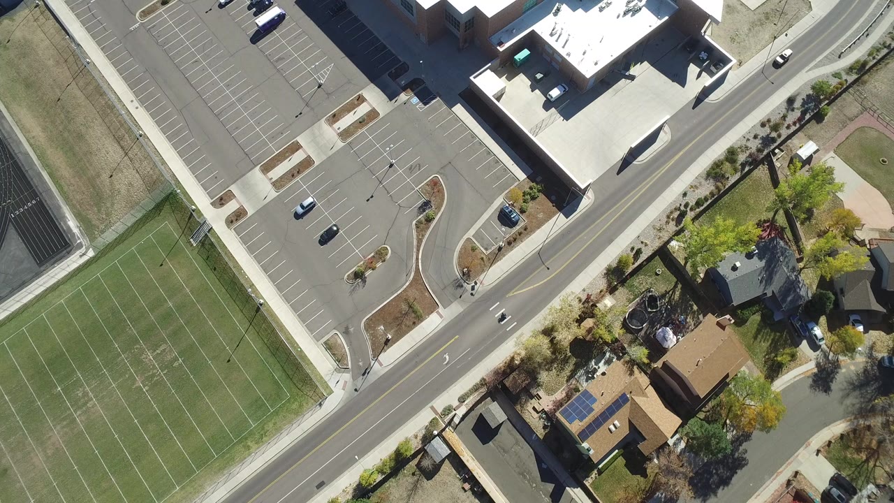 Top-down aerial frame in Hidden Lake, Colorado showing green tennis courts beside a road, a mostly empty parking lot, nearby buildings, trees, homes, and no visible people.