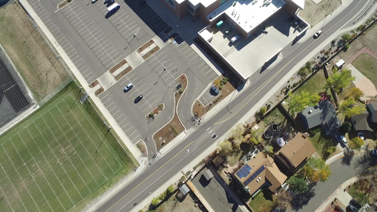 Top-down road scene with tennis courts, a parking lot, buildings, trees, and houses.