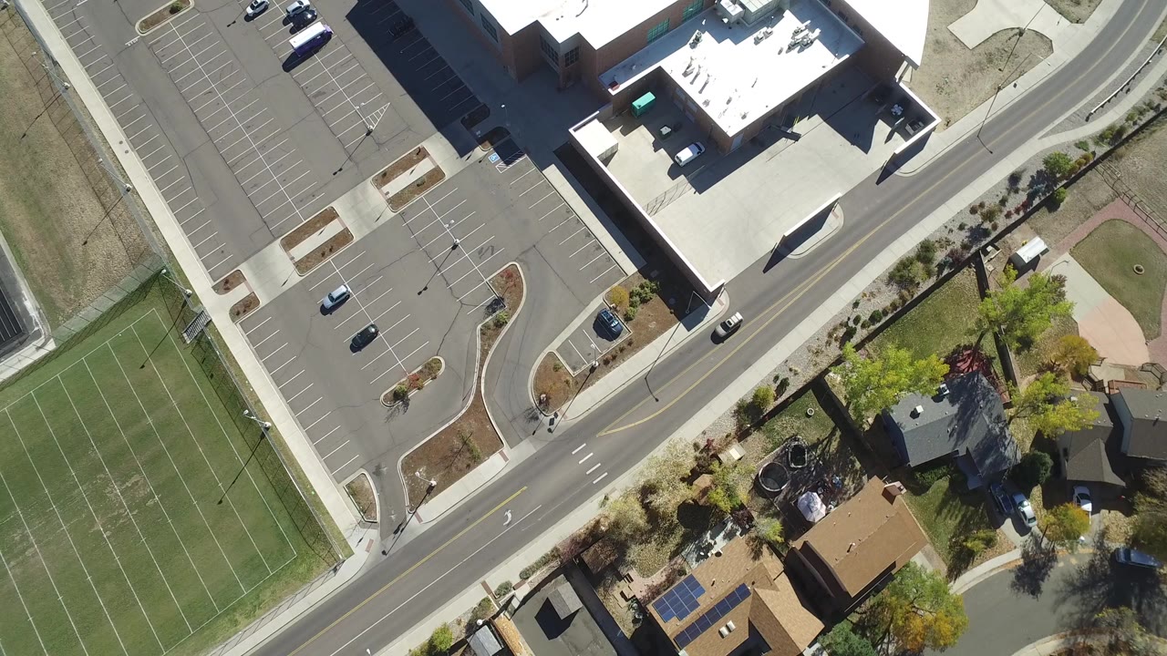 Opening frame over Hidden Lake, Colorado shows a curved road between a large parking lot, marked green sports fields, trees, and private homes, with no visible people.