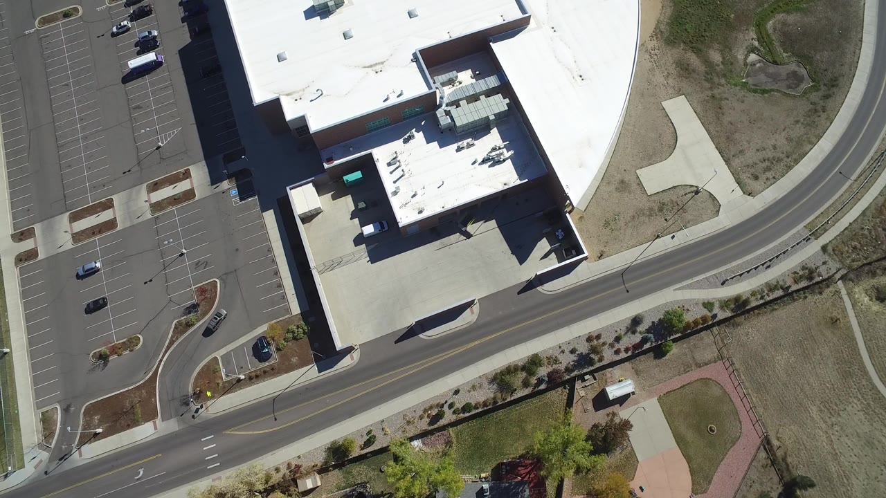 Overhead aerial frame in Hidden Lake, Colorado showing a large white-roofed building beside marked parking lots, a paved road, open dry ground, and no visible people.