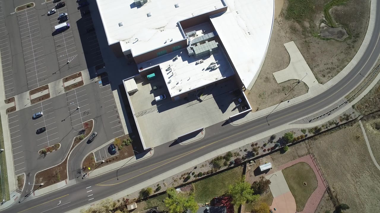 Aerial frame in Hidden Lake, Colorado with the commercial roof shifted slightly across the view, parking rows at left, a curving road below, dry open land at right, and no visible people.