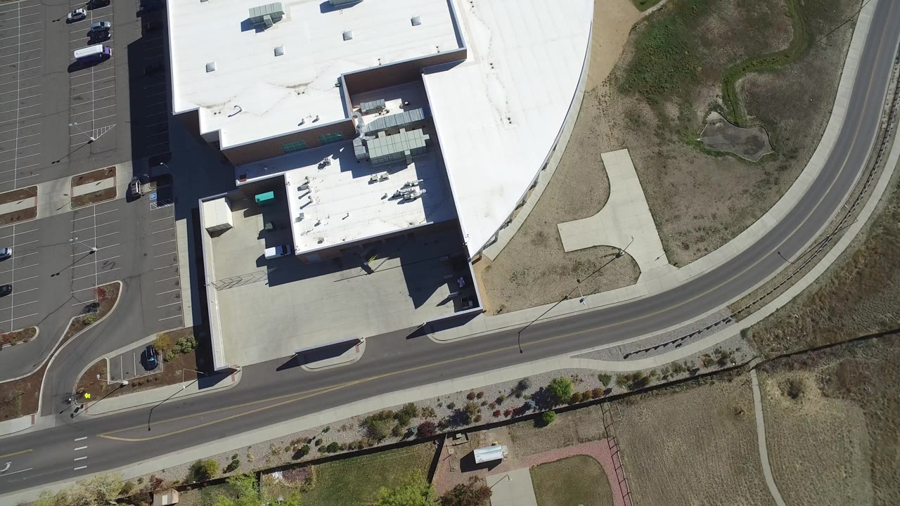 Overhead Hidden Lake, Colorado frame with the large white-roofed building on the left, parking stalls at the edge, a small car on the road, open ground, and no visible people.