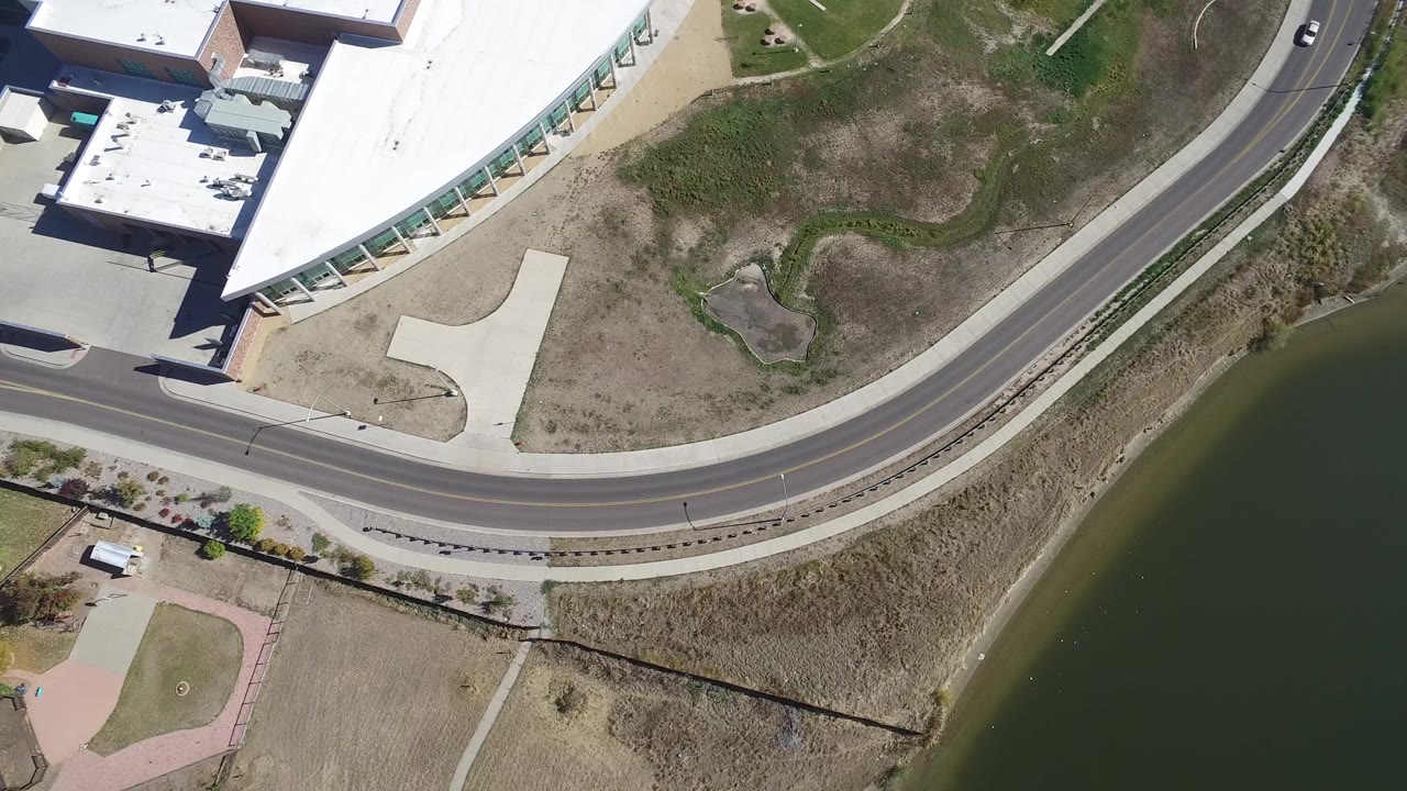 Aerial frame over Hidden Lake, Colorado showing a white-roofed building, curved road, dry open land, lake edge, and no visible people.