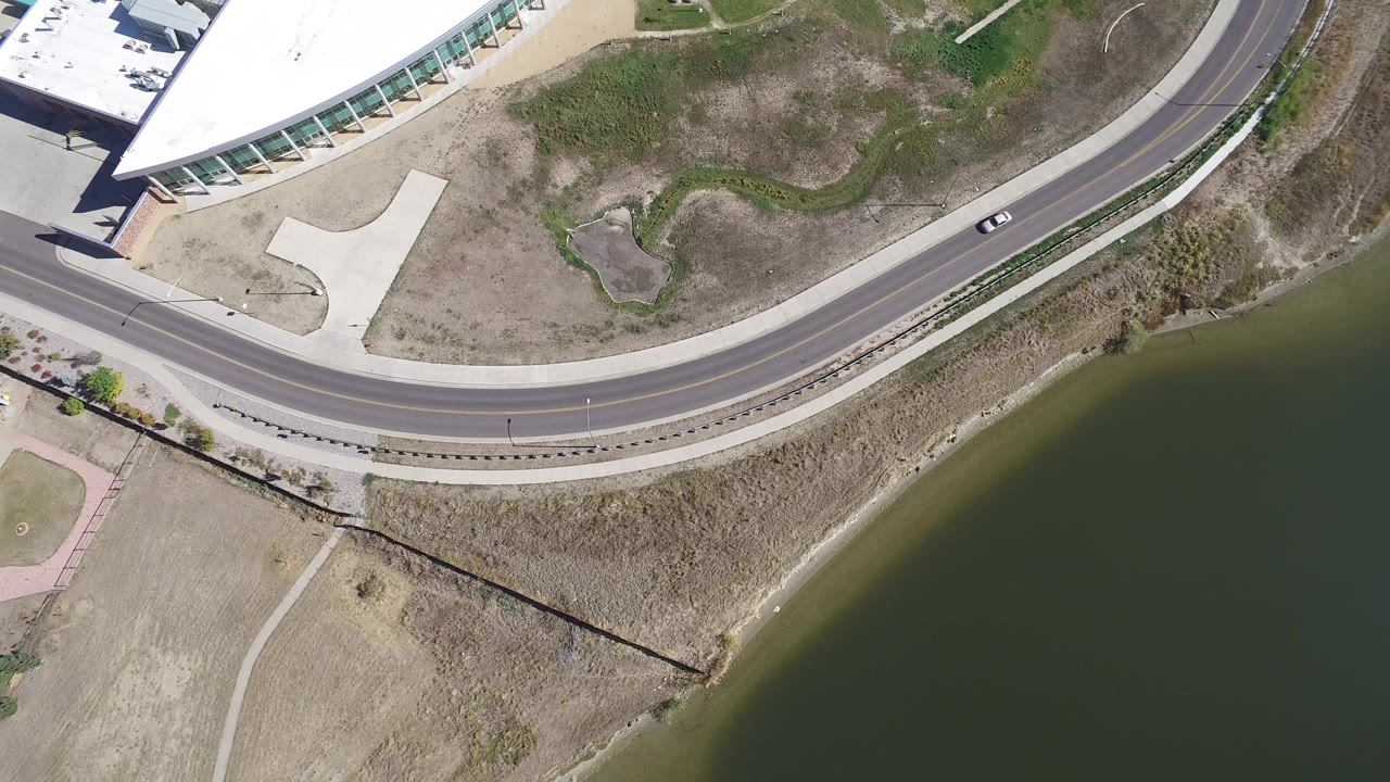 Overhead view of a curved road beside green lake water and dry grass.