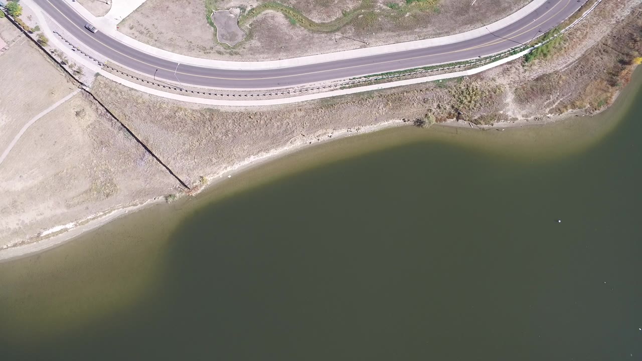 Overhead frame from Hidden Lake, Colorado showing a curved highway above a dry shoreline and olive-green lake water, with no visible people.