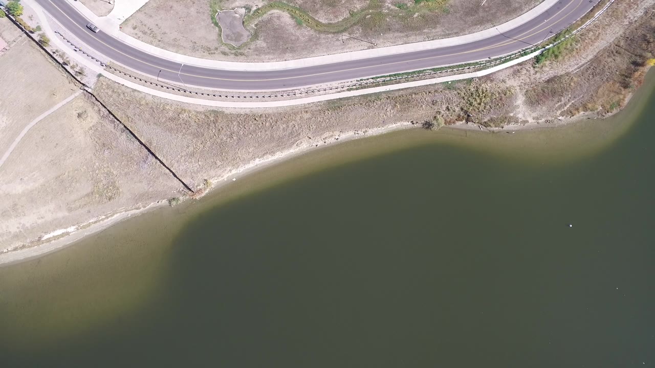 Overhead Hidden Lake, Colorado view with the looping road across the top edge, pale dry land along the bank, and green water filling the lower frame, with no visible people.
