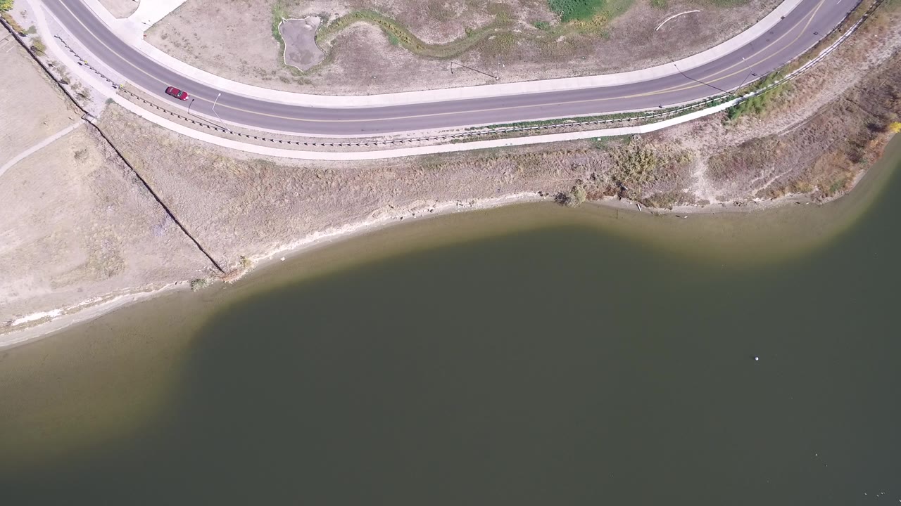 Top-down aerial frame in Hidden Lake, Colorado showing a curving road, a red car near the upper left, dry grass, shoreline, dark water, and no visible people.