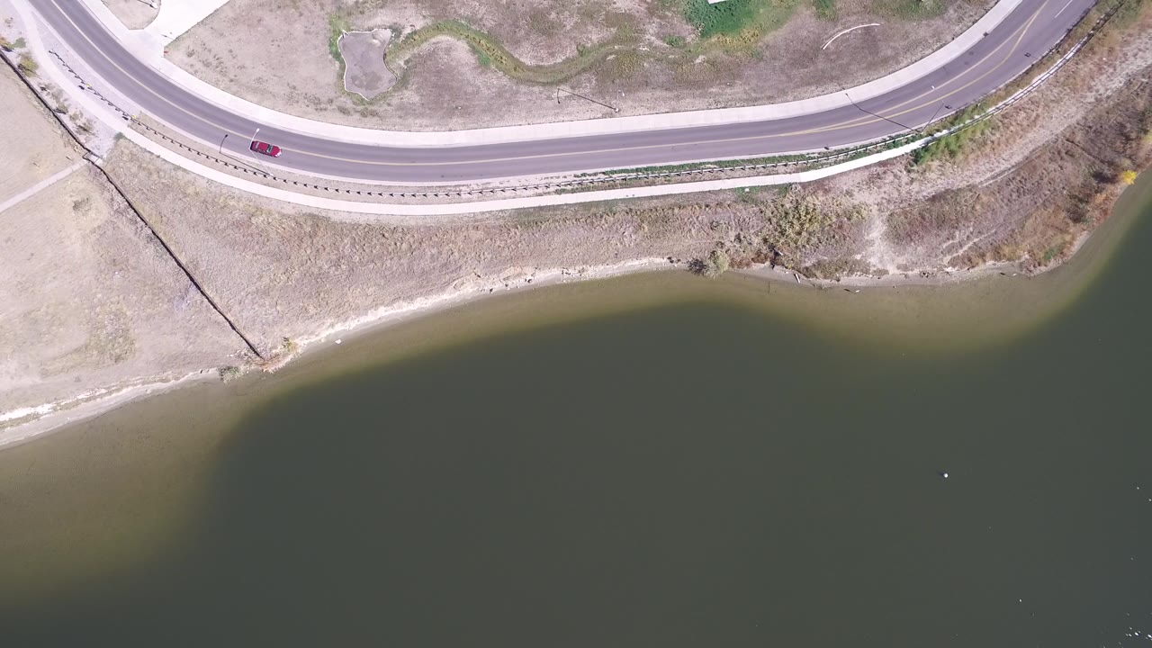 Straight-down view of a lakeside road with dry grass and open water.