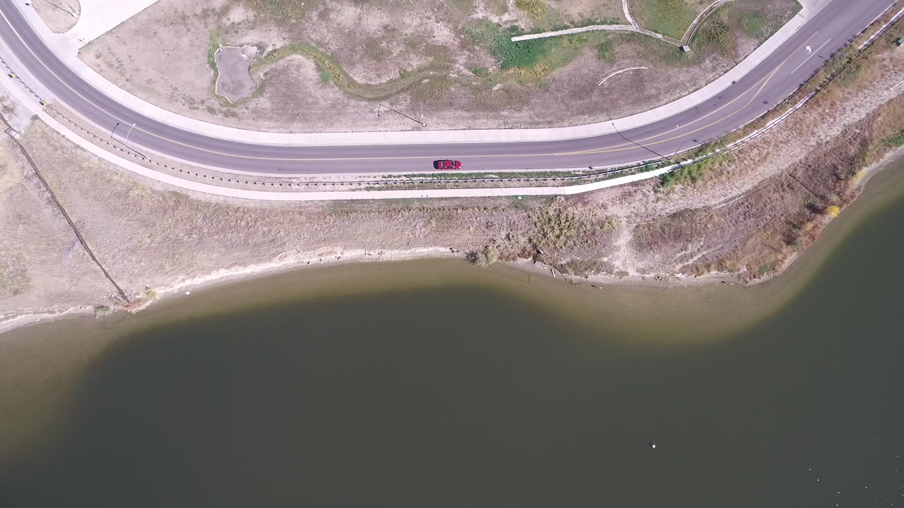 Overhead aerial frame of Hidden Lake, Colorado with green water below, pale shoreline, dry scrub land, a curved road above, and no visible people.