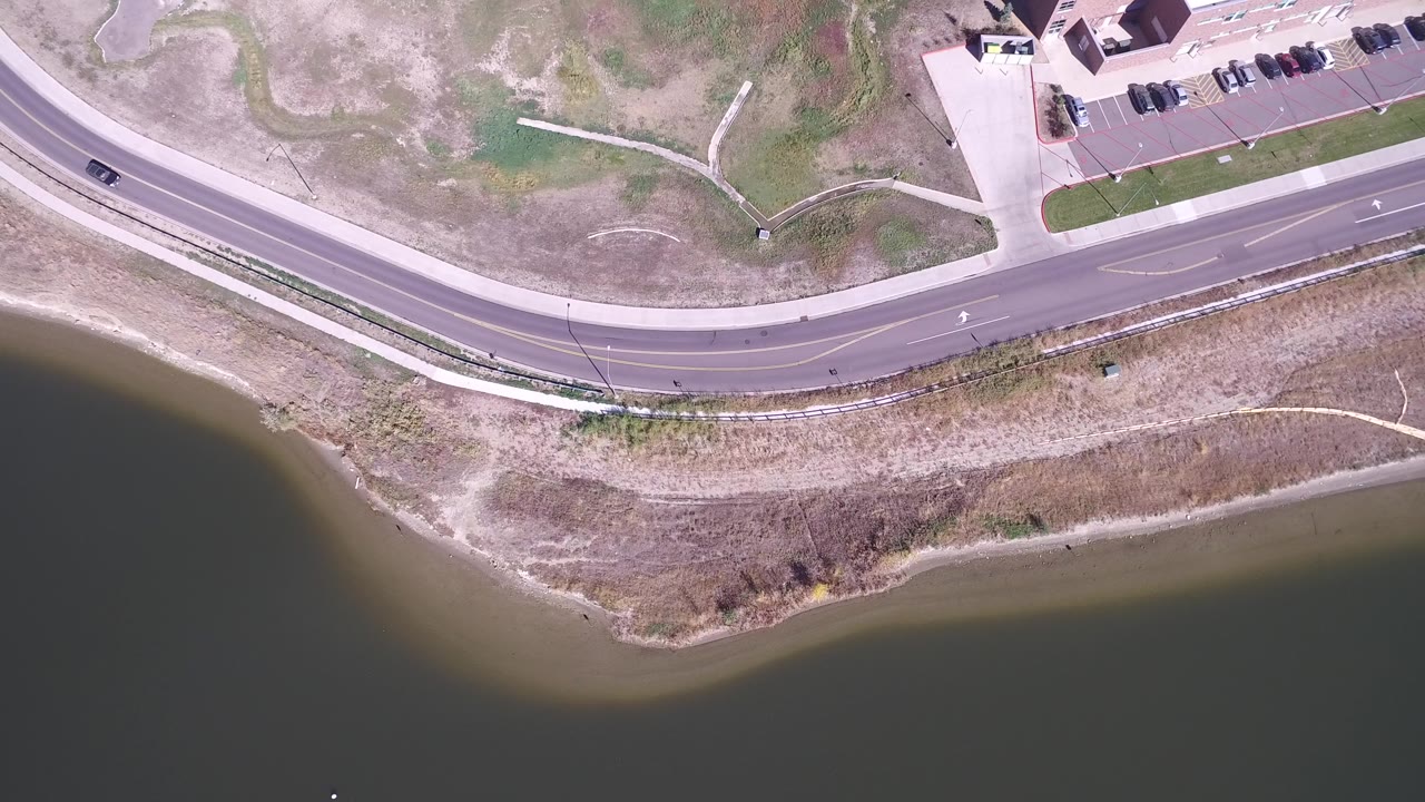 Overhead view of a curved road beside lake water and dry grass.
