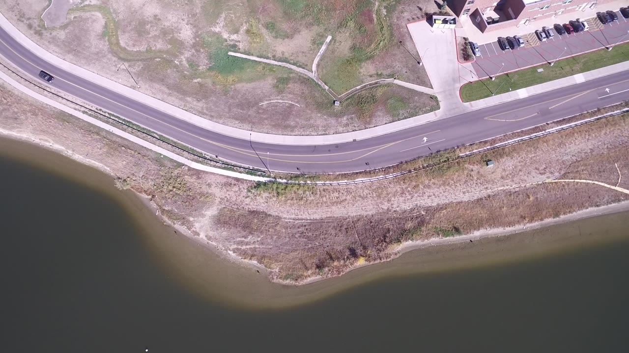 Top-down view in Hidden Lake, Colorado showing the curved road, a dark car near the upper lanes, lake water below, dry grass strips, and no visible people.