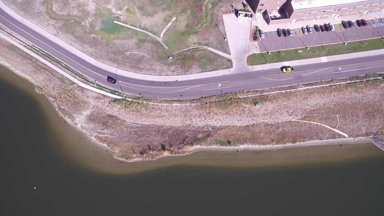 Overhead view of a lakeside road, shoreline, parking lot, and cars.