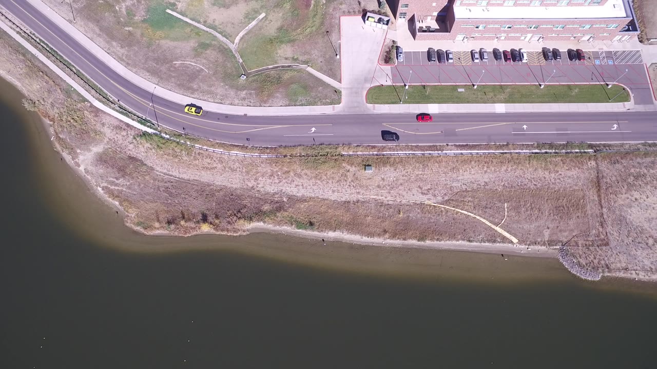 Aerial view of cars on a road beside lake water and a parking lot.