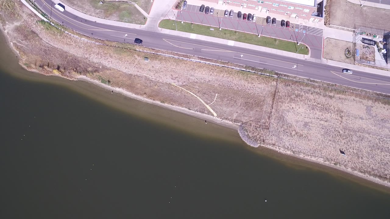 Hidden Lake, Colorado appears from above with the lake across the lower frame, dry shoreline in the middle, cars on the road above, and small distant pedestrians along the path area.