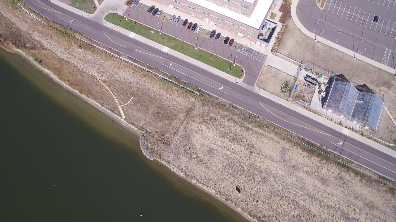 Overhead frame of Hidden Lake, Colorado showing dark lake water, pale shoreline grass, a two-lane road, parked cars, and a glass-roofed building with no visible people.