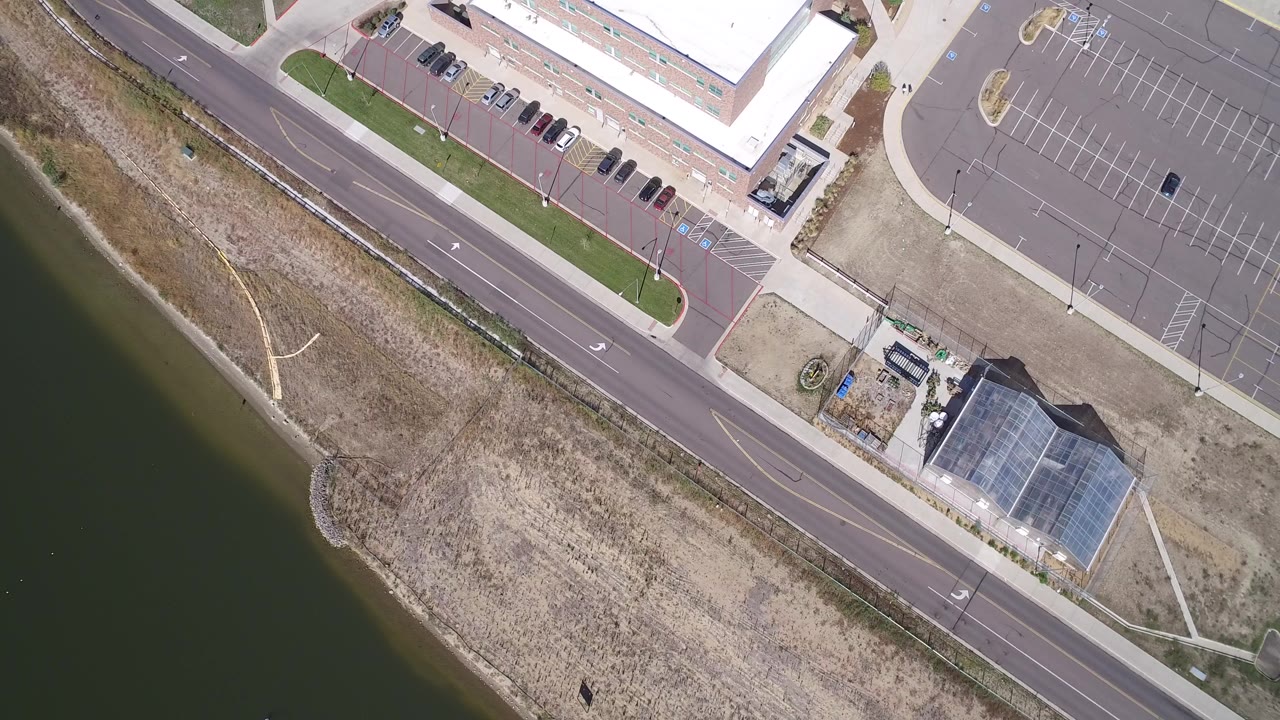 Overhead frame from Hidden Lake, Colorado showing dark lake water beside dry shoreline grass, a diagonal road, a mostly empty parking lot, and small buildings with no visible people.