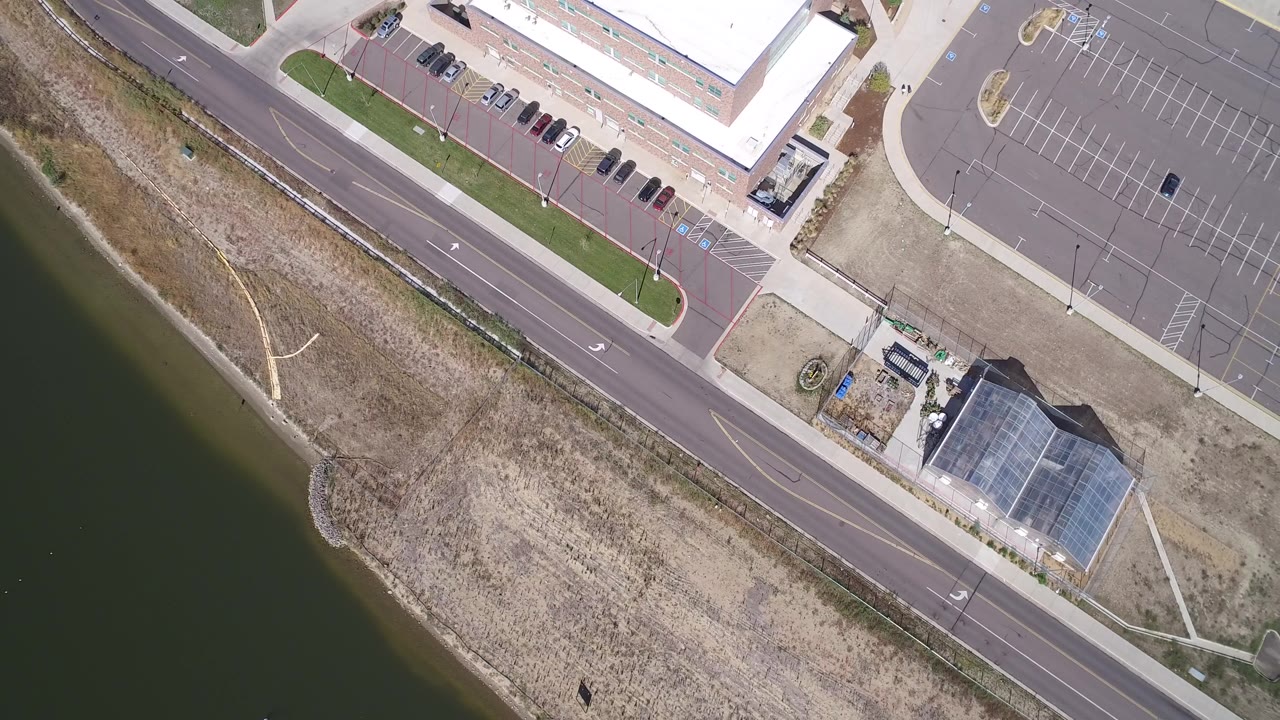 High overhead frame from Hidden Lake, Colorado with the road cutting between the lake shoreline and a large empty parking lot, plus small buildings near the lower right and no visible people.