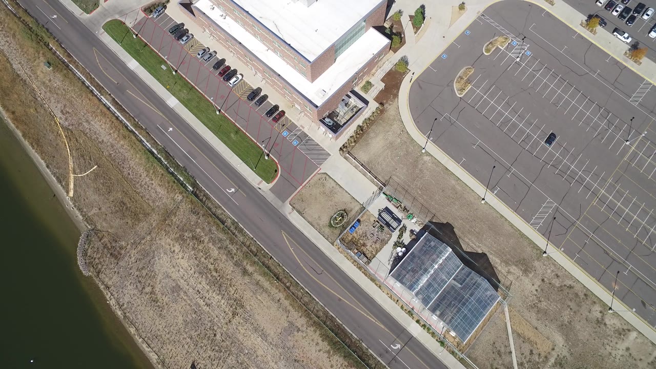 Overhead aerial frame in Hidden Lake, Colorado showing a road beside dry ground, a mostly empty commercial parking lot, a white-roofed building, and no visible people.