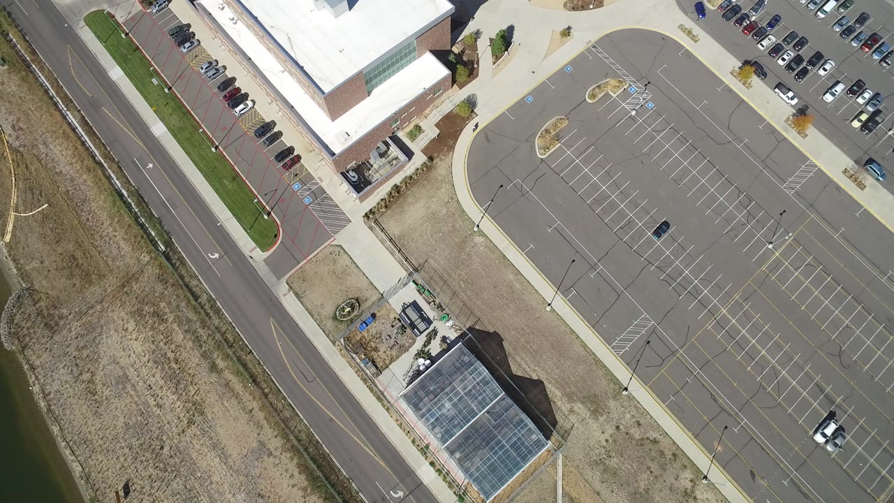 At 0:00, an overhead view in Hidden Lake, Colorado shows a large mostly empty school parking lot, building roofs, a bordering road, dry grass, parked cars, and no visible people.