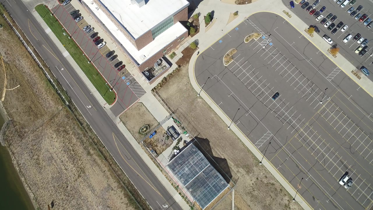 At 0:01, the Hidden Lake aerial frame centers the marked parking rows with school buildings at the top, a road at the left, dry grass nearby, and no visible people.