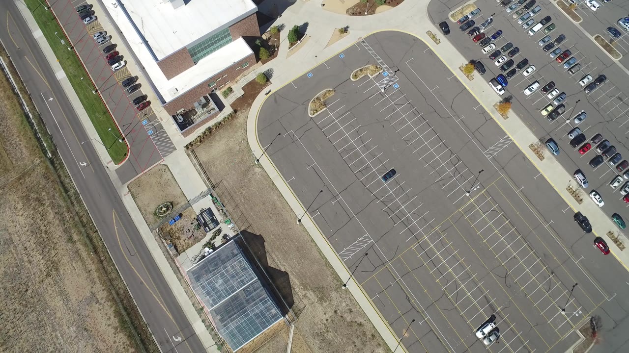 Opening overhead frame of the Heston Town Center parking lot in Hidden Lake, Colorado, with a road at left, commercial buildings at top and lower left, parked cars at right, and no visible people.