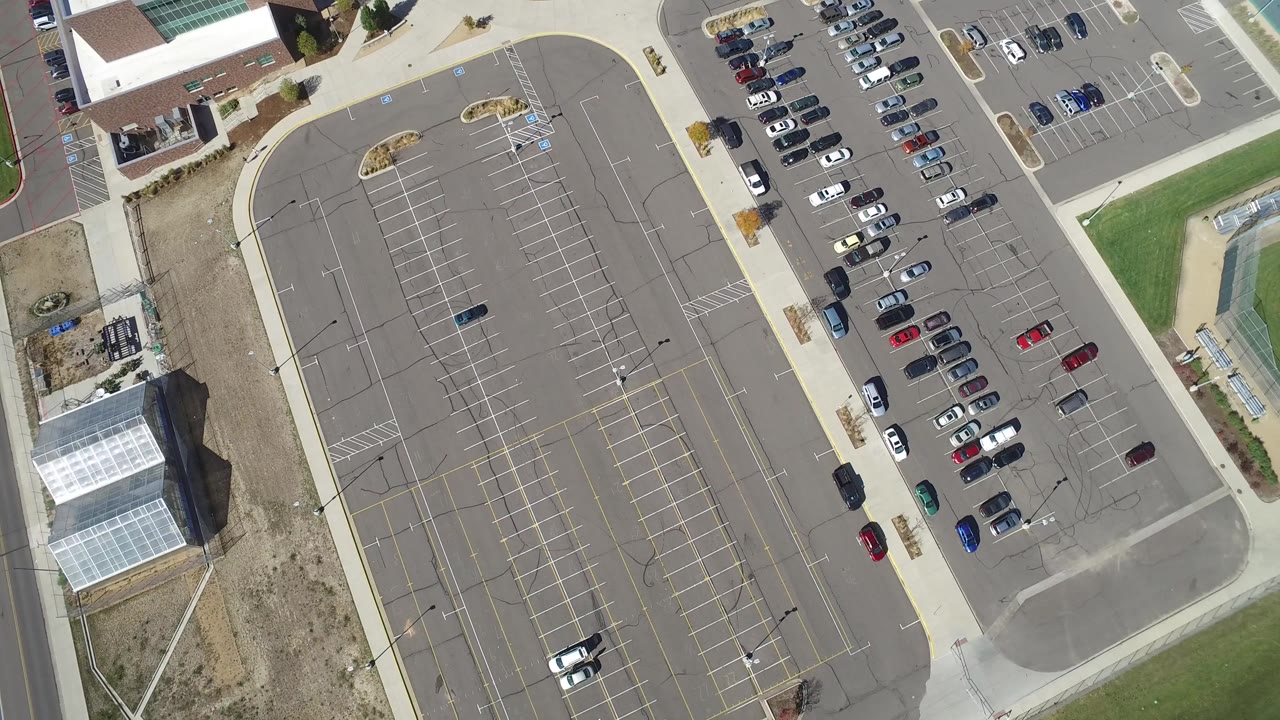 Overhead parking lot with painted rows and scattered cars beside buildings.