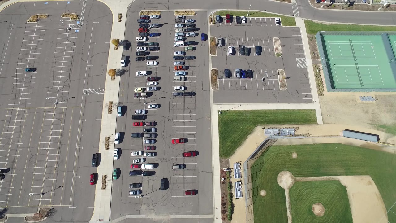 Overhead Hidden Lake, Colorado frame with clustered parked cars on the left, green tennis courts at upper right, paved sidewalks, and a baseball field along the lower edge with small pedestrians present.