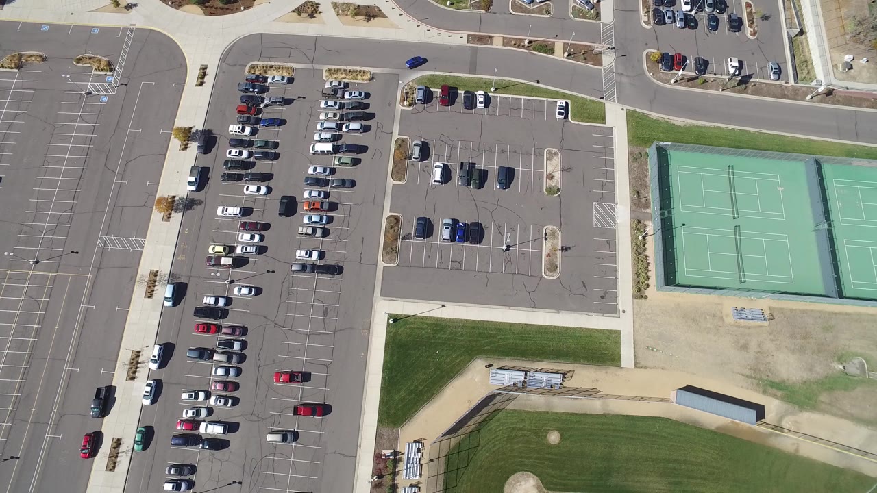 Top-down aerial frame from Hidden Lake, Colorado showing organized parked cars, curved access roads, green tennis courts, sidewalks, grass, dirt field space, and no visible people.