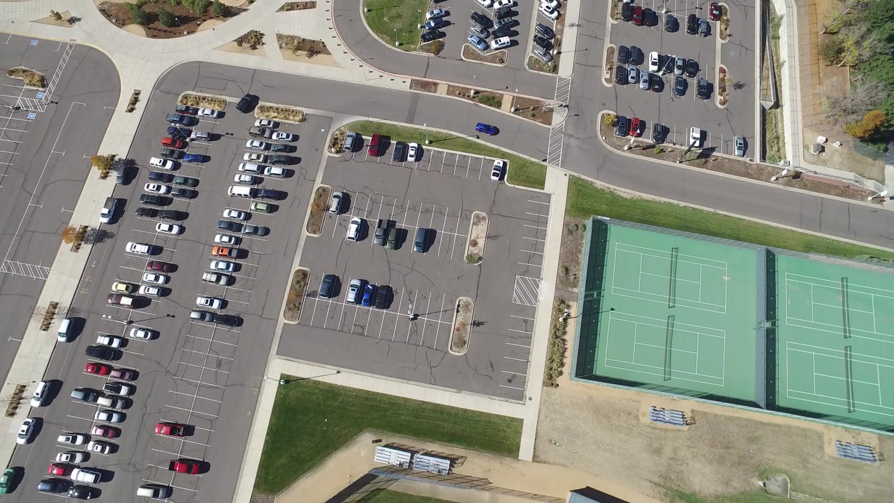 Overhead frame in Hidden Lake, Colorado showing rows of parked cars, empty spaces, roads, sidewalks, grass, and two green tennis courts, with no visible people.