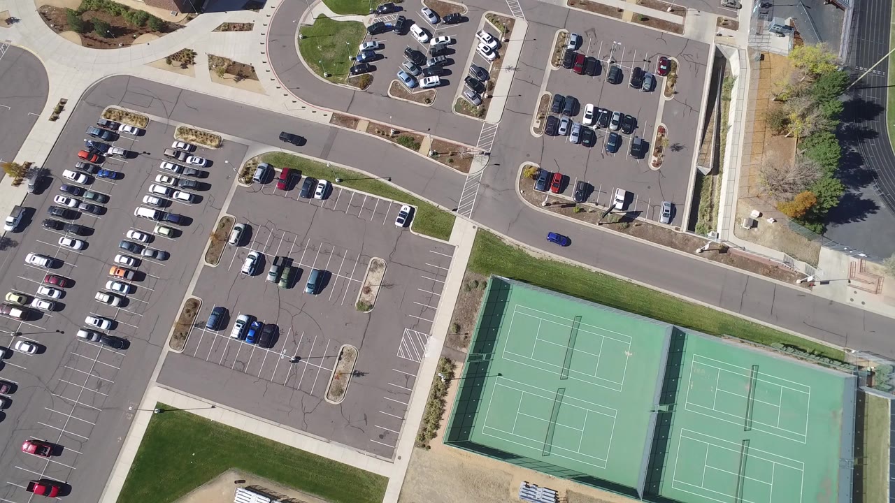 Opening overhead frame in Hidden Lake, Colorado showing parking rows, a road, green tennis courts, sidewalks, grass, trees, and no visible people.