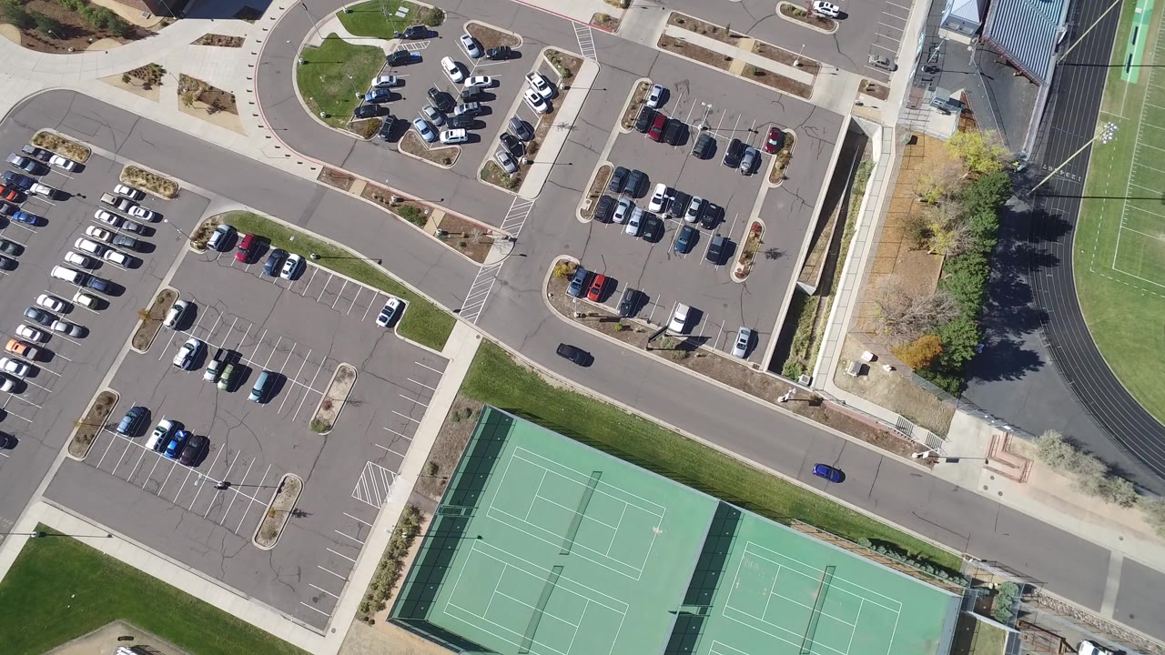 A high overhead frame in Hidden Lake, Colorado shows parked cars, a road, green tennis courts, sidewalks, and the edge of a sports field, with no clearly visible people.