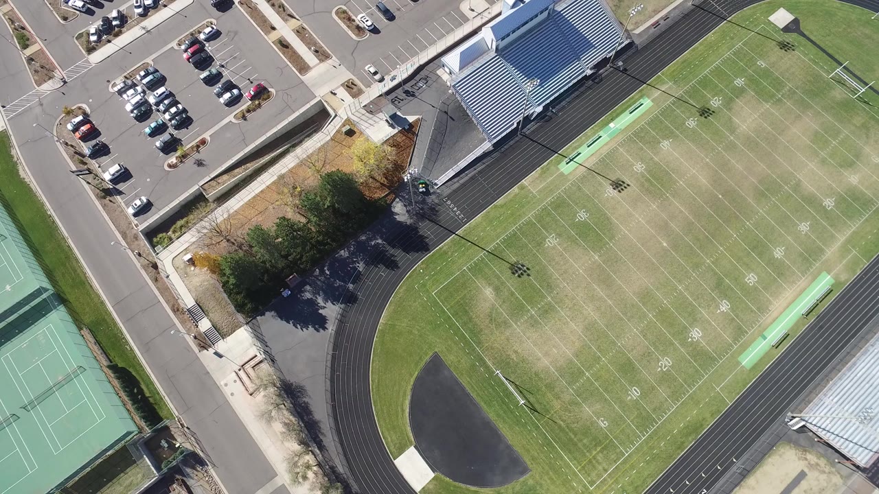 Overhead view of a turf sports field, running track, tennis courts, bleachers, roads, and parking.