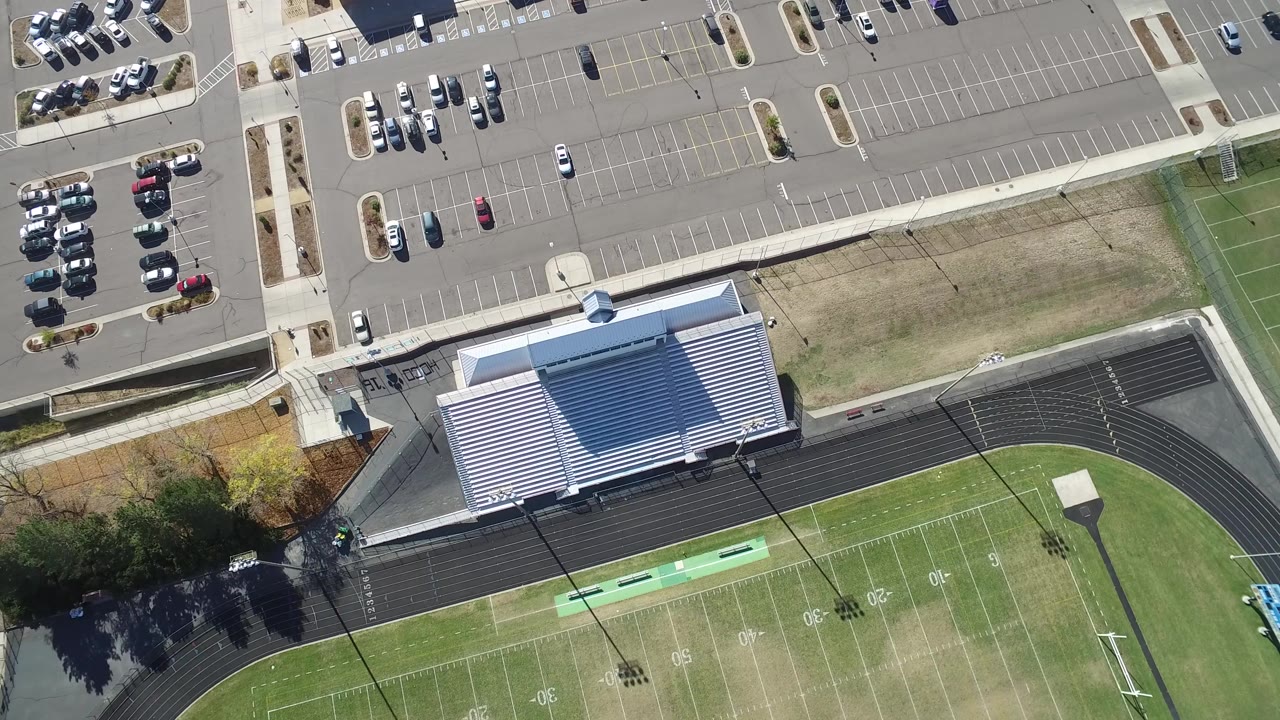 Overhead view in Hidden Lake, Colorado showing the football field edge, black running track, metal bleachers, sidewalks, parked cars, and no visible people at the start of the clip.