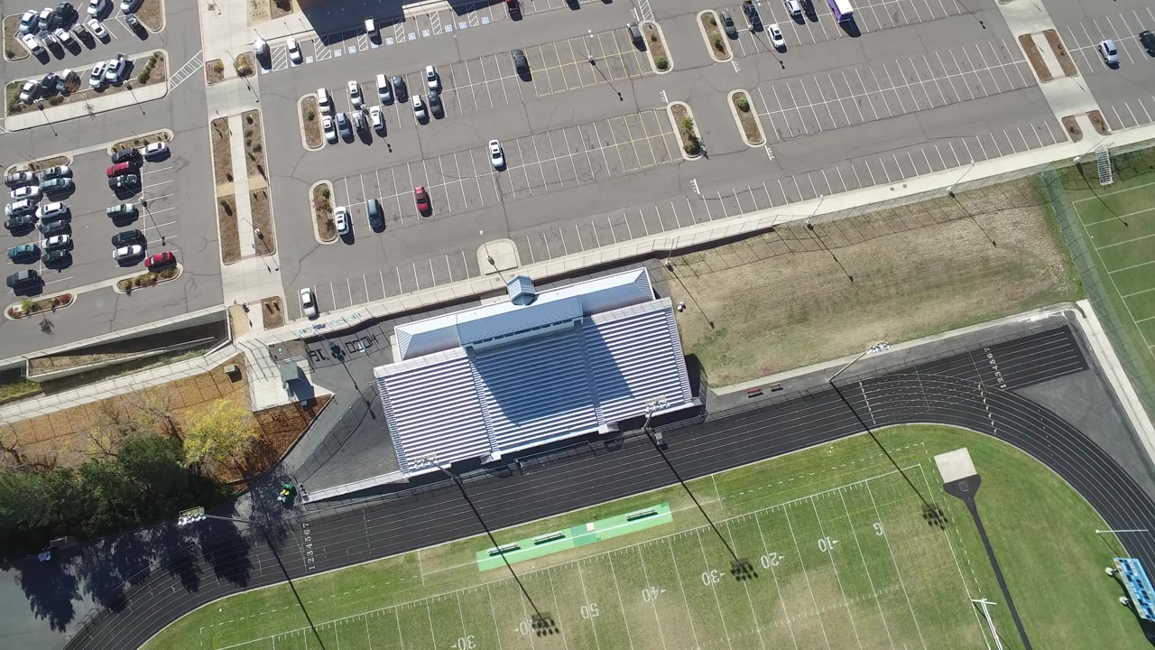 Top-down sports field with bleachers, track, and parked cars.