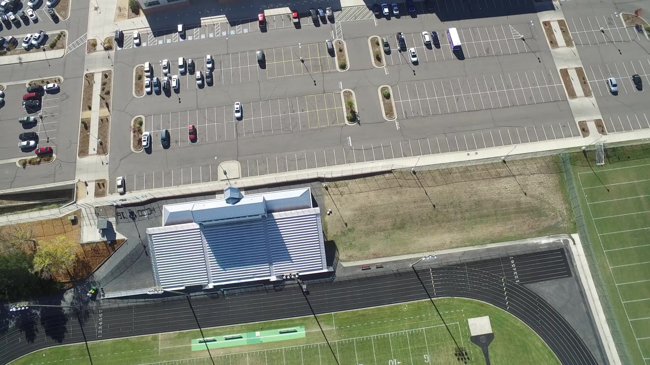 High aerial frame over Hidden Lake, Colorado showing a mostly empty parking lot above a metal-roofed building, a running track, and green field turf, with no visible people.