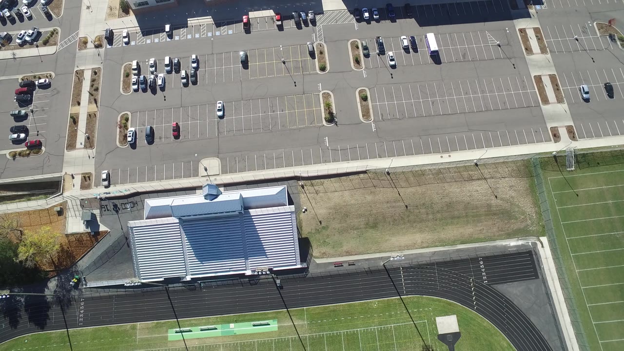 Top-down parking lot with sidewalks, parked cars, building roof, and athletic field.