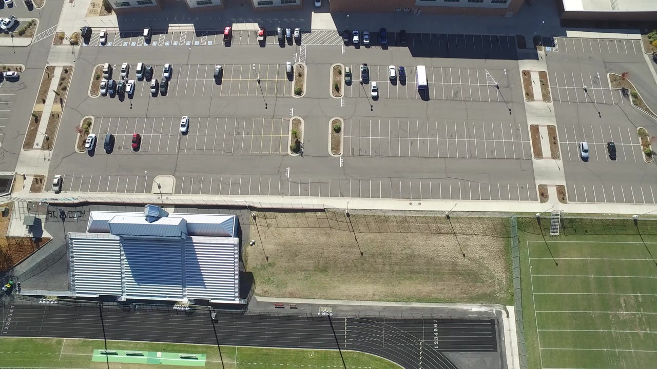 Opening overhead frame of a Hidden Lake, Colorado parking lot with scattered cars, a large roofed building, turf fields, a running track, and no visible people.