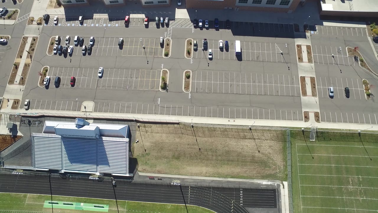 Suburban parking lot with parked cars, sidewalks, and green field edges.