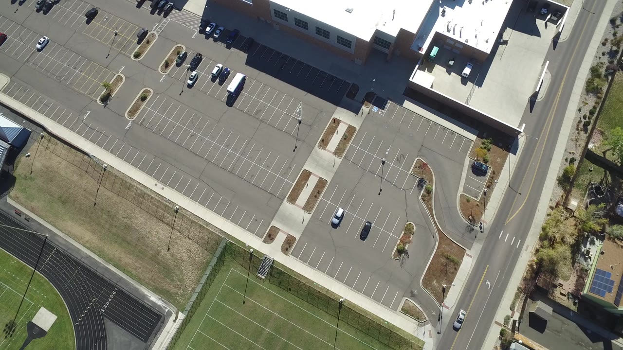 Opening overhead frame in Hidden Lake, Colorado showing a large building edge, mostly empty parking lot, vertical road, sports field corner, nearby houses, trees, and no visible people.