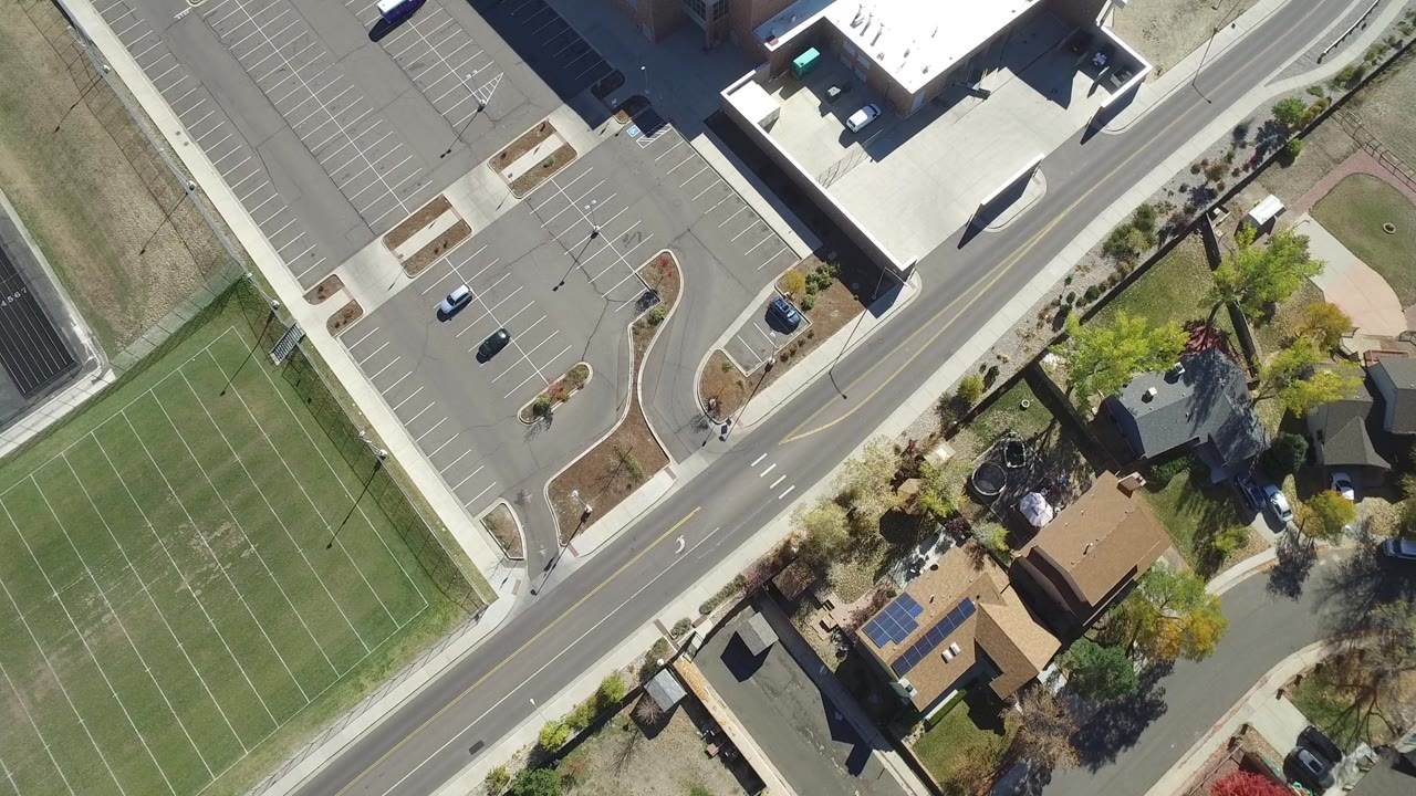 Top-down aerial frame in Hidden Lake, Colorado showing a diagonal road between a green sports court, a parking lot, commercial buildings, trees, and homes, with no visible people.