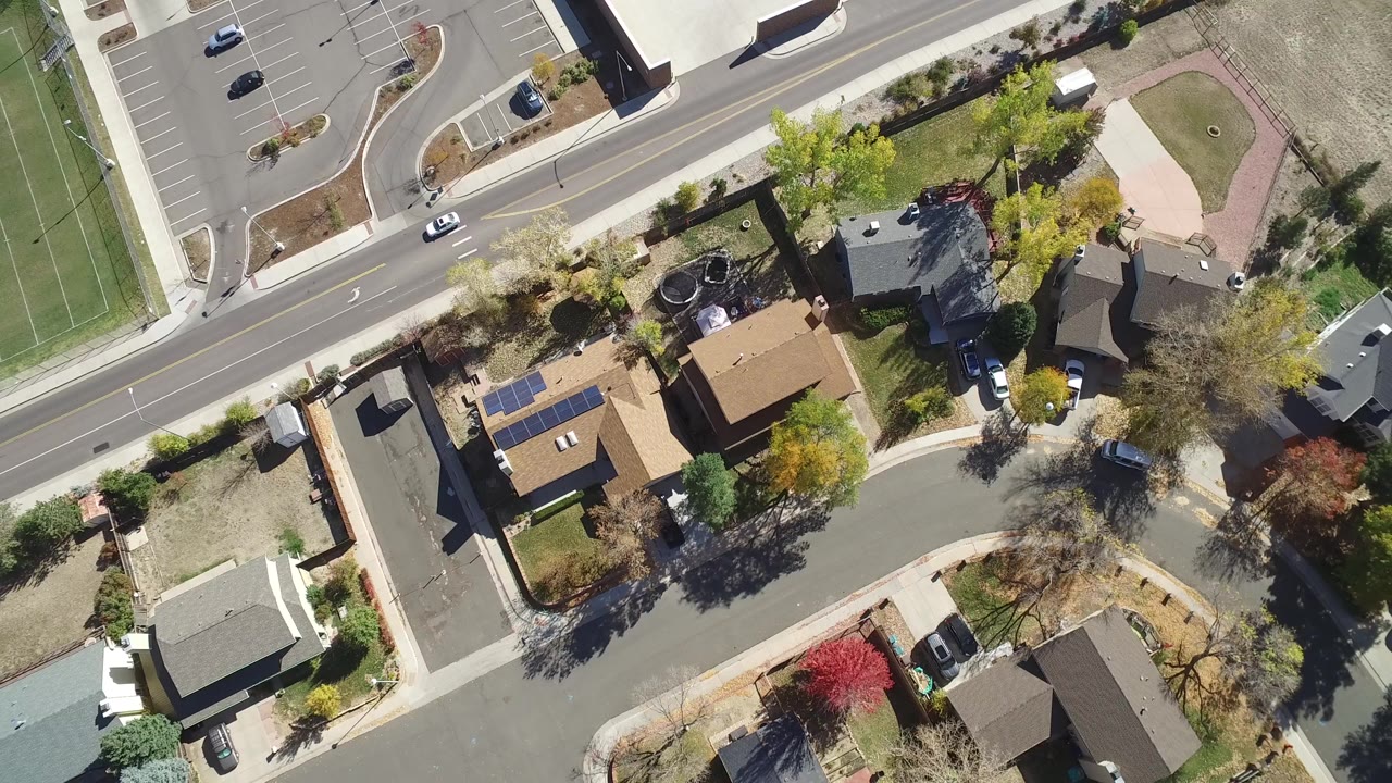 Overhead frame from Hidden Lake, Colorado showing houses, a curving residential street, a larger road, parked cars, trees, and the edge of a green sports field, with no visible people.