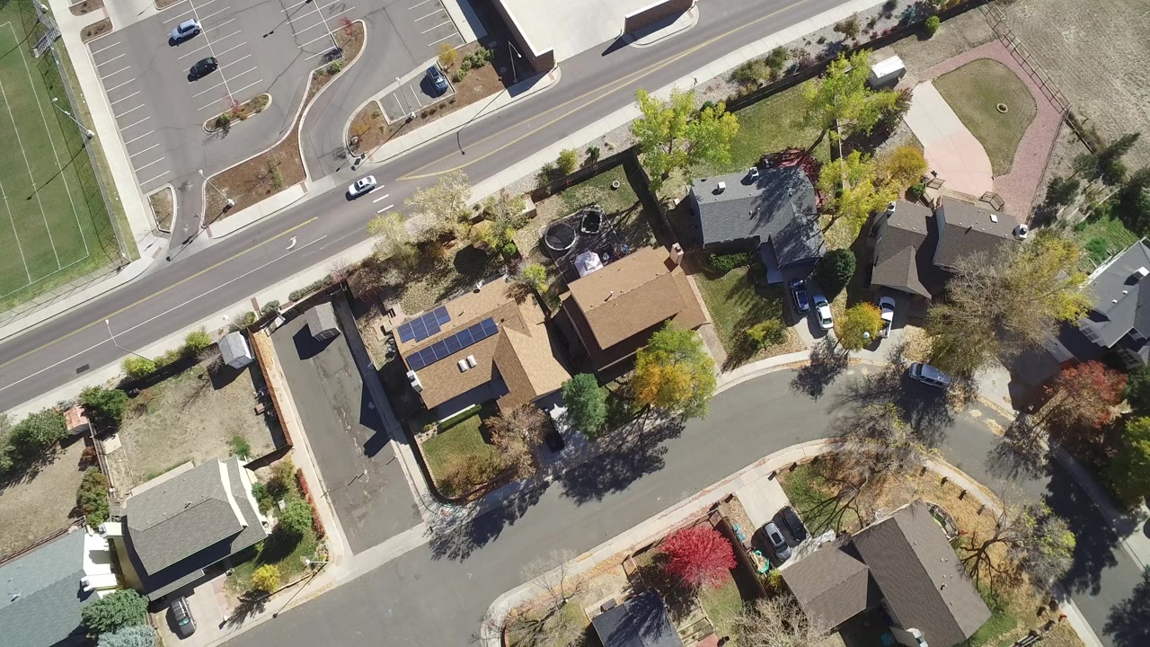 Aerial frame from Hidden Lake, Colorado with several private homes, fenced yards, a curved street, parked vehicles, sidewalks, and mixed green and autumn trees, with no visible people.
