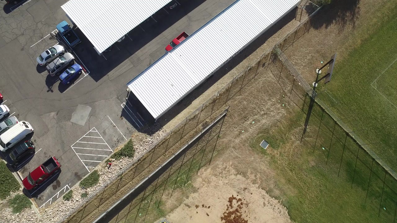 Aerial frame in Hidden Lake, Colorado with two long white parking covers, vehicles on the paved side, dry grass between fences, green field markings, and no visible people.