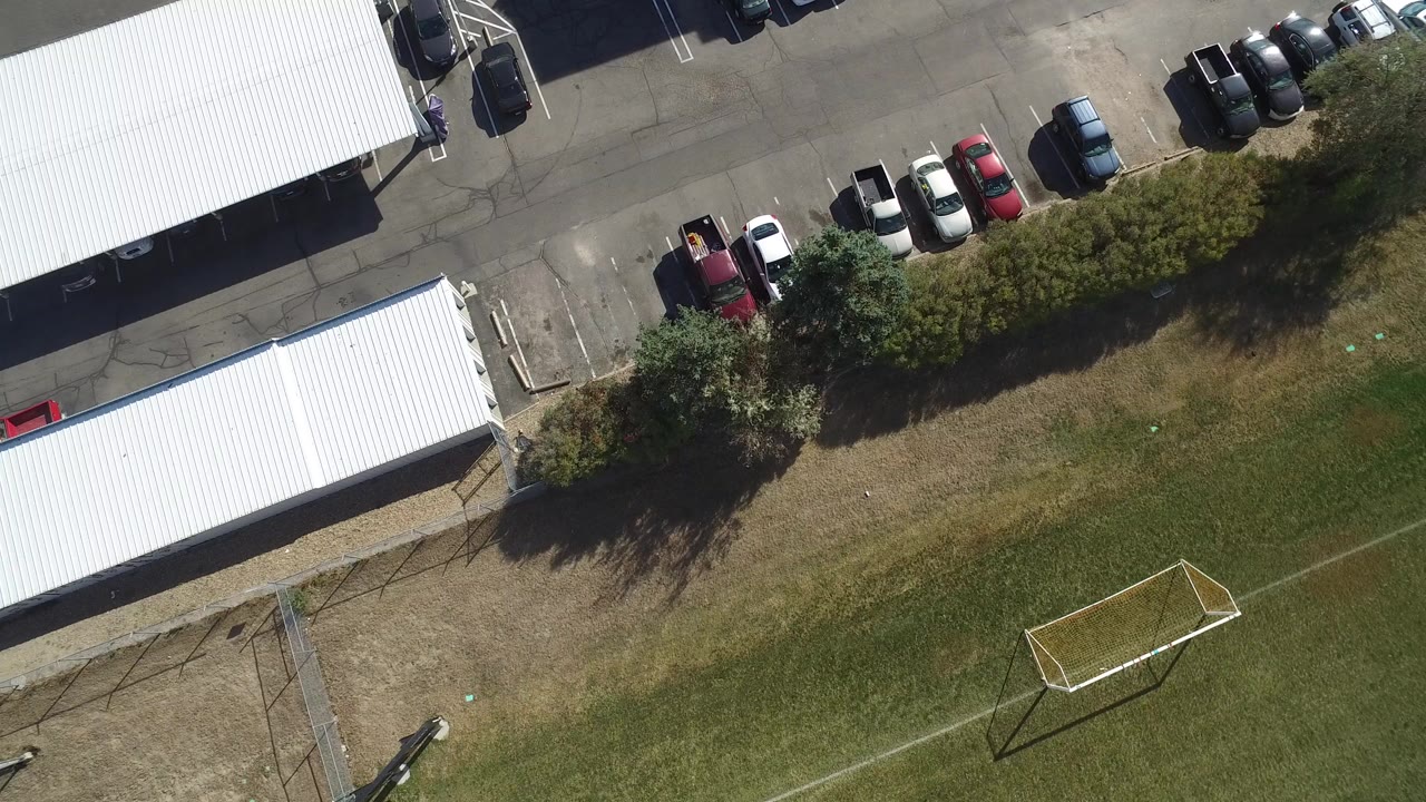 Overhead view of a soccer field edge, parking lot, trees, and white-roofed buildings.