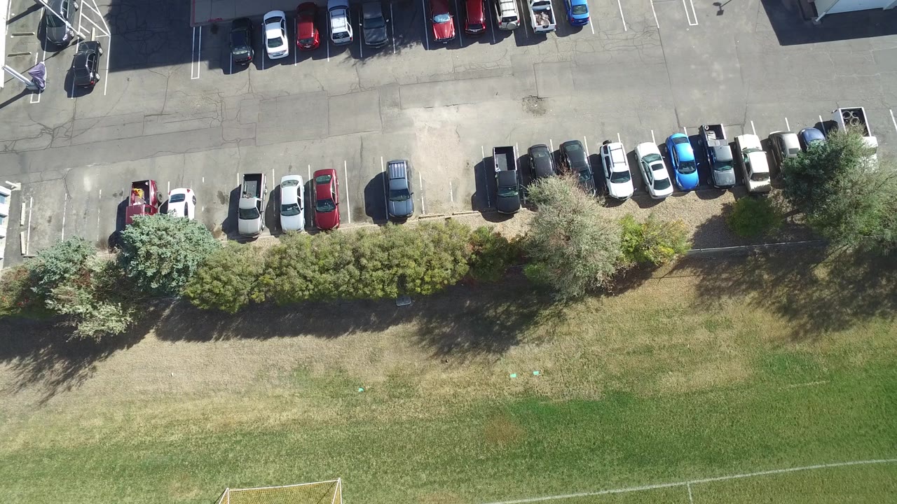 Near-vertical aerial frame in Hidden Lake, Colorado, showing parked cars along an asphalt lane, dense shrubs, open grass, and a small person visible near the upper roadway area.