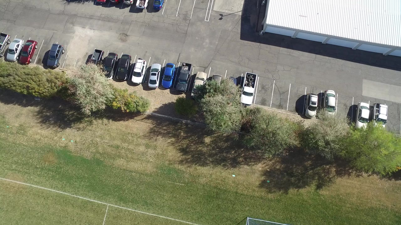 Opening overhead frame in Hidden Lake, Colorado showing parked cars along a back lot, a light-roofed building, trees, grass, and no visible people.