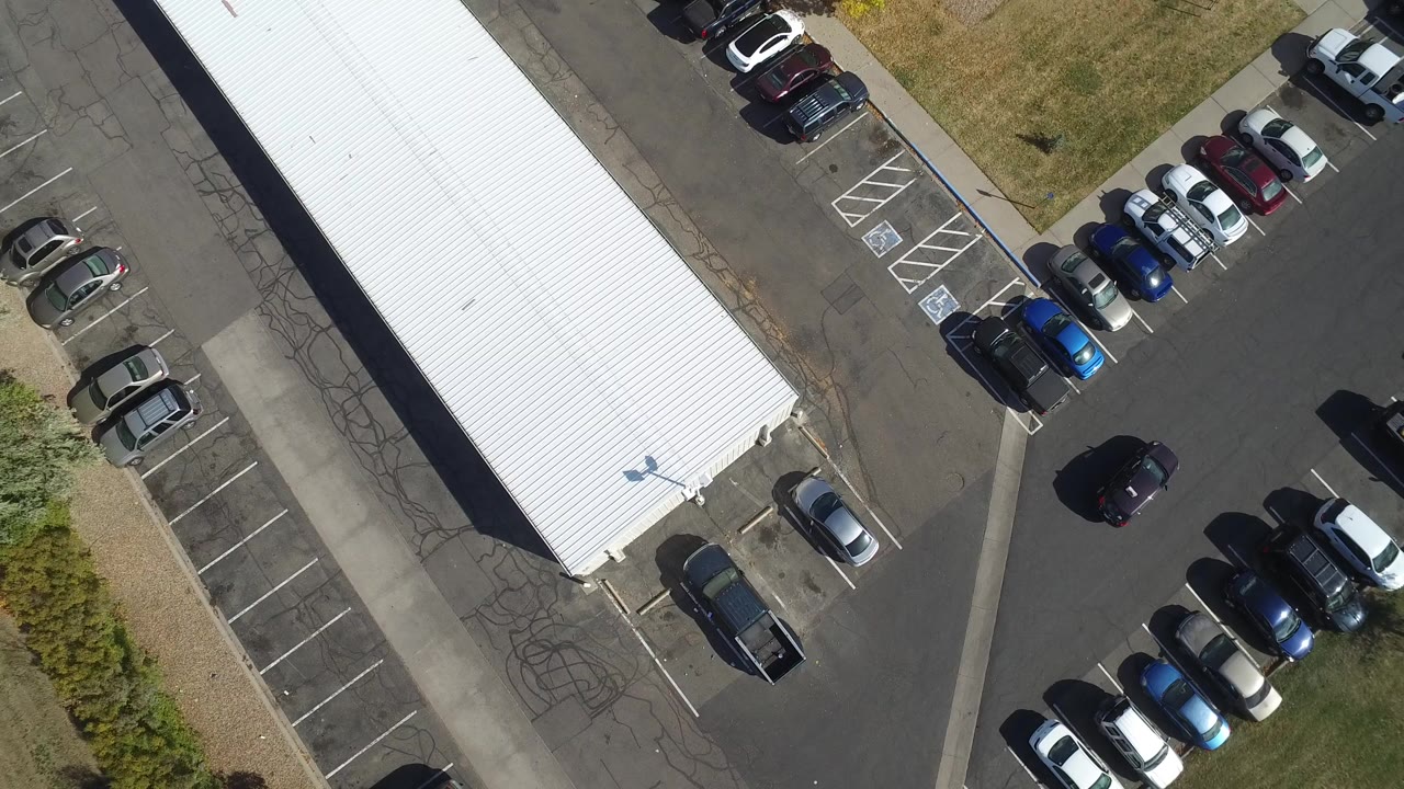 Overhead frame in Hidden Lake, Colorado shows the edge of a white-roofed building, rows of parked cars, asphalt lanes, grass, and no visible people at the start of the clip.